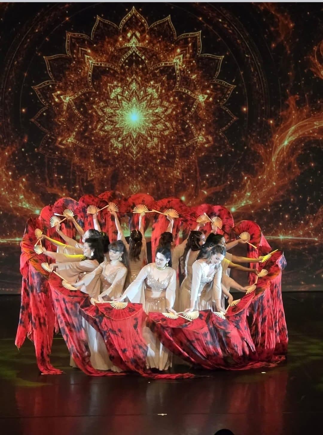 Danseuses de Surya en cercle, éventails de soie rouge déployés devant un mandala lumineux, lors du spectacle de fin d'année